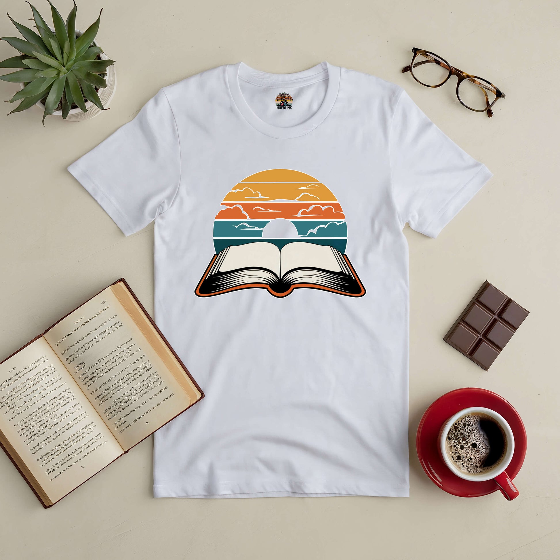 White tee with open book under a colorful sky design, symbolizing vibrant literary journeys. Paired with books, coffee, and glasses.