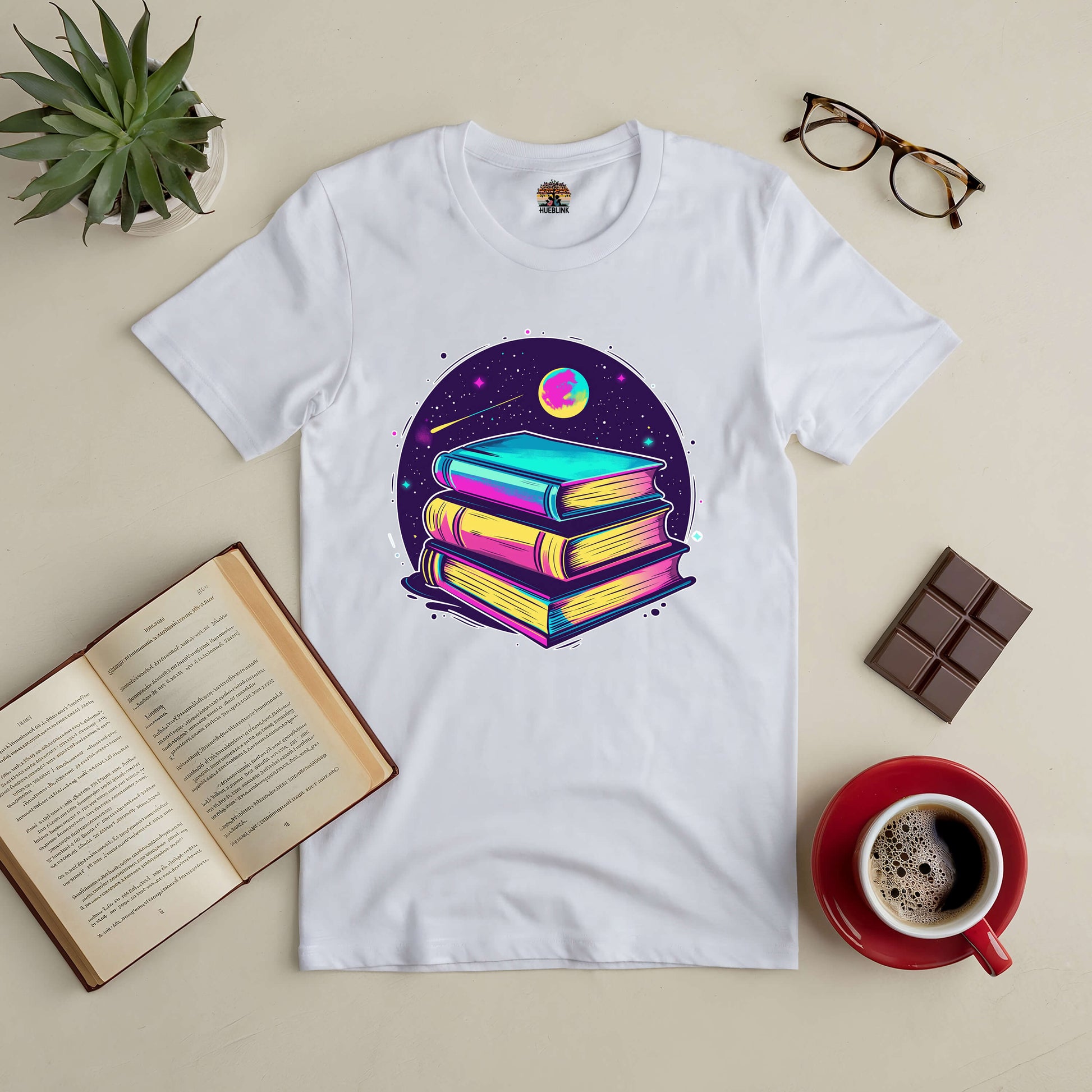 White tee with vibrant books and galaxy design, surrounded by coffee, books, and chocolate. Perfect for book and space lovers.