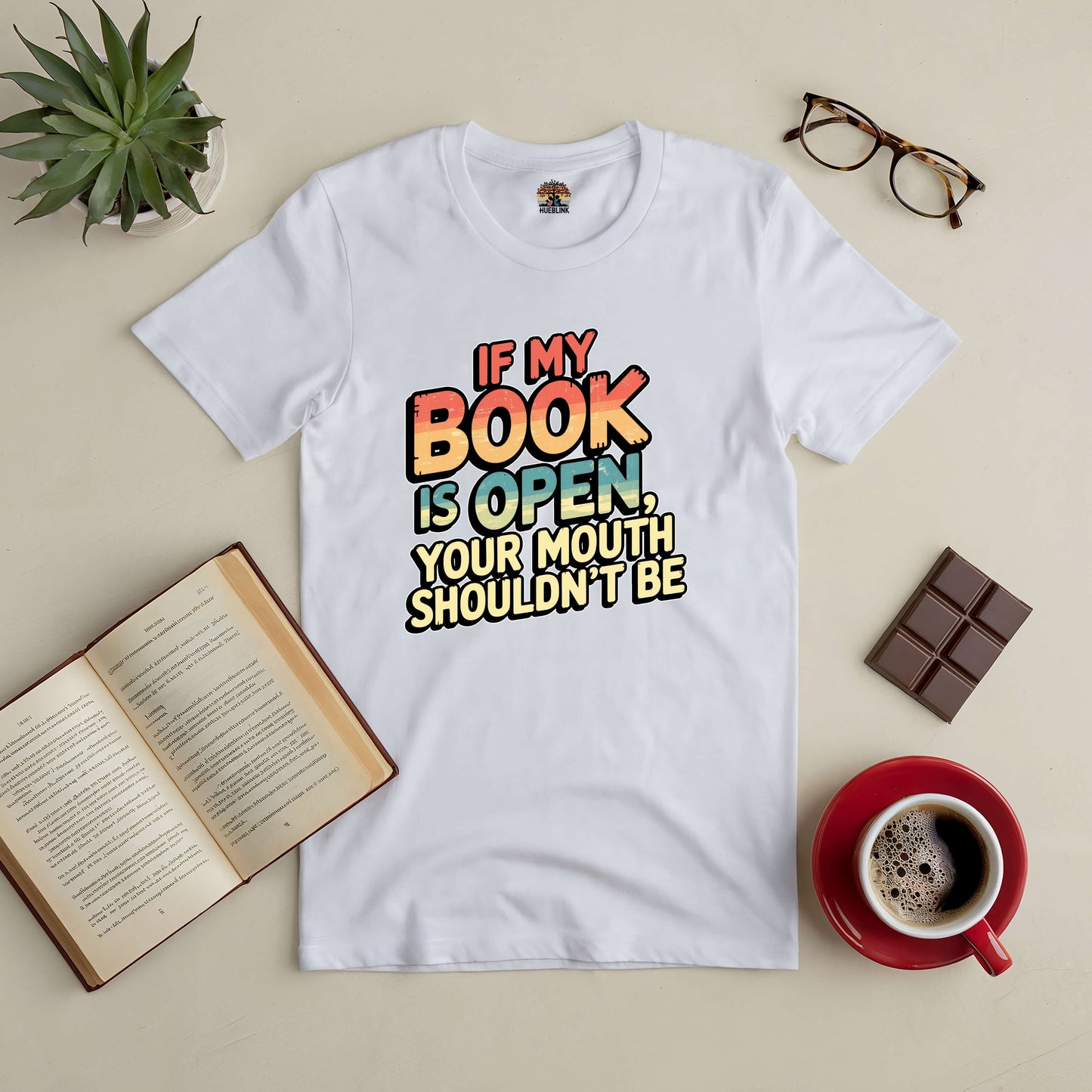 White tee with "If My Book Is Open, Your Mouth Shouldn't Be" text, surrounded by a book, coffee, glasses, and a plant. Perfect for book lovers.