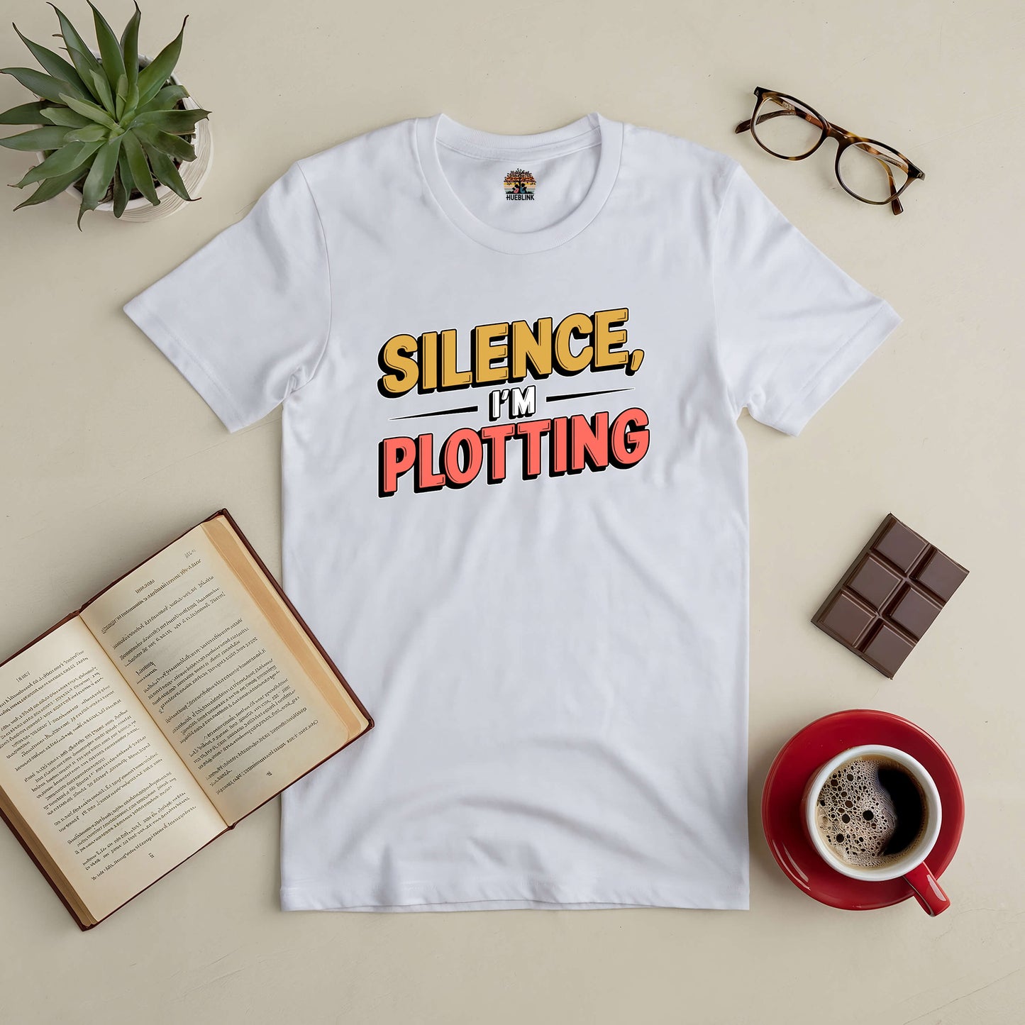 White tee with bold text "Silence, I'm Plotting" beside a book, coffee, and glasses, perfect for writers and book lovers.