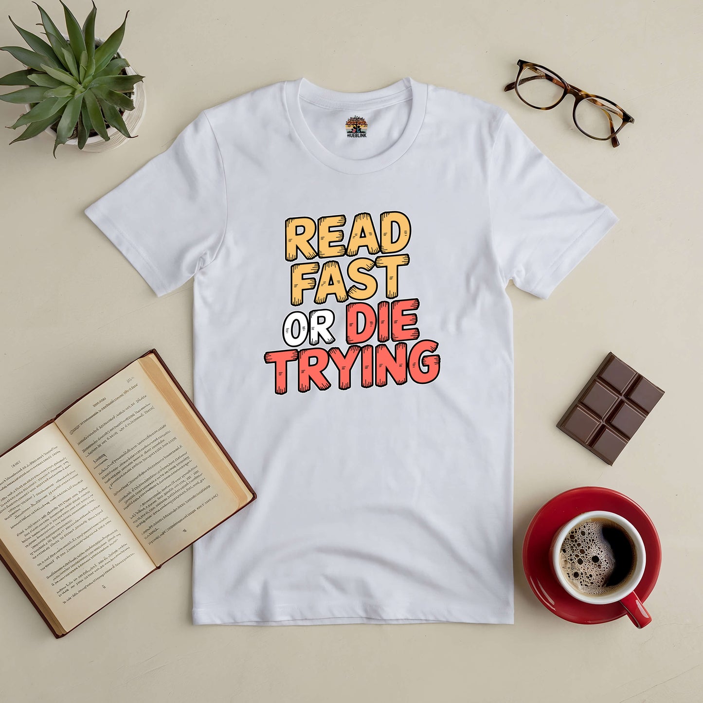 White "Read Fast or Die Trying" tee surrounded by book, coffee, chocolate, glasses, and plant for book lovers.